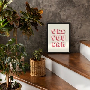 Może przedstawiać: Wpisany w ramę motywacyjny plakat z tekstem "YES YOU CAN" w r&oacute;żowych literach z białym obramowaniem na białym tle. Plakat wisi na ścianie z drewnianymi schodami na pierwszym planie.