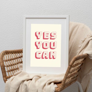 Może przedstawiać: Biały wydruk w ramie z tekstem "YES YOU CAN" w pogrubionych, r&oacute;żowych literach z efektem 3D. Wydruk leży na wiklinowym krześle z białym kocem.