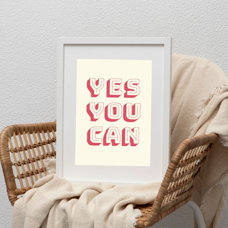 Może przedstawiać: Biały wydruk w ramie z tekstem "YES YOU CAN" w pogrubionych, r&oacute;żowych literach z efektem 3D. Wydruk leży na wiklinowym krześle z białym kocem.