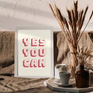 Może przedstawiać: Biały wydruk w ramie z tekstem "YES YOU CAN" w kolorze r&oacute;żowym z białym obramowaniem. Wydruk leży na brązowej powierzchni w kratkę z wazą suszonego pszenicy i białym ceramicznym czajnikiem.