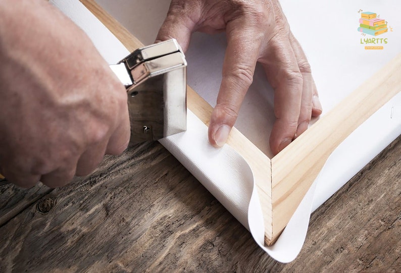 May include: A person using a staple gun to stretch white canvas over a wooden frame. The frame is made of light-colored wood, and the canvas is being secured at the corner. The background is a rustic wooden surface.