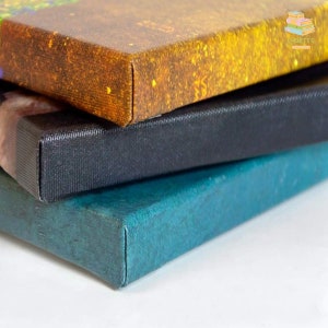 May include: Close-up of three stacked canvas art prints. The top print has a textured, golden-yellow surface. The middle print is black, and the bottom print is teal. The edges of the canvases are neatly folded and stapled.