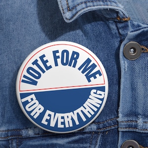以下が含まれることがあります： 赤、白、青のカラーリングで「Vote for me for everything」と書かれた白と青のボタン。ボタンは青いデニムジャケットにピンで留められています。