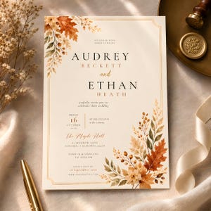 May include: A rectangular wedding invitation card featuring a cream background with a thin gold border. The text reads Audrey Beckett and Ethan Heath, with autumn-themed watercolor illustrations of orange and brown maple leaves and berries.