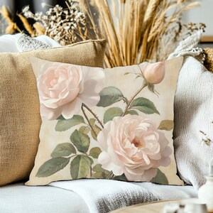 May include: A square throw pillow featuring a botanical illustration of two blooming pale pink roses and a single closed rosebud. The floral design includes detailed green leaves and stems set against a soft, cream-colored background.