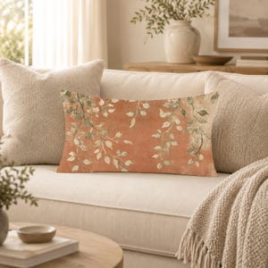 May include: A rectangular throw pillow featuring a terracotta orange background with a repeating pattern of delicate, light green trailing vine and leaf illustrations. The pillow has a soft, textured fabric appearance and a rectangular shape.