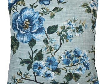 Könnte beinhalten: Quadratisches Zierkissen mit hellblauem Hintergrund und floralem Muster. Das Design zeigt große, detaillierte blaue Blumen und kleinere weiße und blaue Blüten mit grünen Blättern und Stielen. Das Kissen hat ein strukturiertes Aussehen.