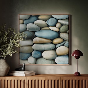 Puede incluir: Una obra de arte enmarcada que presenta un primer plano de piedras lisas y coloridas en tonos de azul, beige y verde. La obra de arte se exhibe sobre un mueble de madera con un jarrón de vegetación, un pájaro decorativo, libros y una lámpara burdeos.
