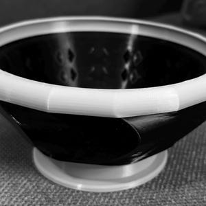 May include: A black and white bowl with a geometric design. The bowl has a white rim and base, and the interior is black. The bowl is sitting on a gray surface.