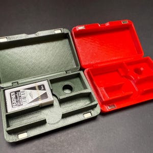 May include: An open, olive green case with a red case next to it. The olive green case contains a package of razor blades. The red case is empty. Both cases have magnetic closures. The razor blade package has black and white text.