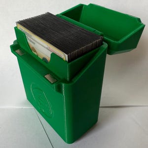 May include: A bright green, 3D-printed card storage box with a hinged lid. The box is open, revealing a stack of trading cards. The box has a magnetic closure and a circular design on the front. The cards are labeled "Trainer" and "Supporter".