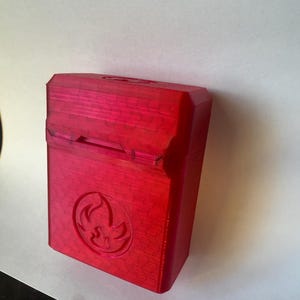 May include: A red rectangular box with a hinged lid. The box has a textured surface and a circular emblem with a flame design. The box is made of a solid material and is likely 3D-printed.