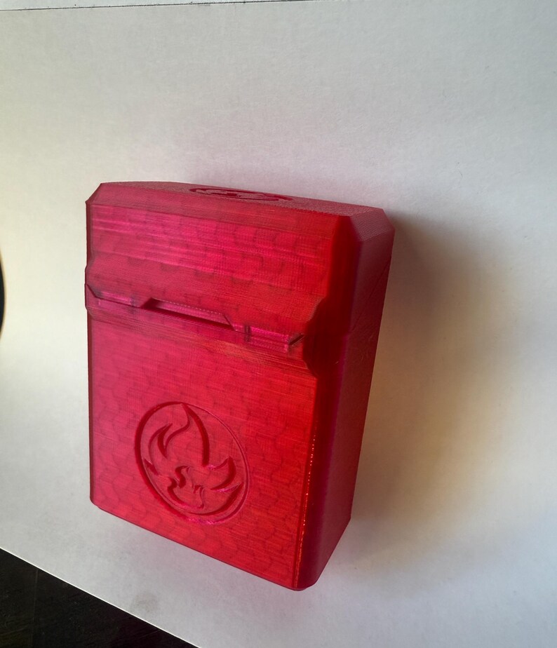 May include: A red rectangular box with a hinged lid. The box has a textured surface and a circular emblem with a flame design. The box is made of a solid material and is likely 3D-printed.