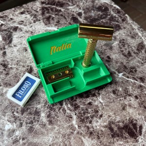 以下が含まれることがあります： 金色の安全カミソリと緑色のケース。ケースは開いており、カミソリの刃と金色の文字で「Italia」と書かれています。「Filoso」の替刃パックがケースの隣にあります。