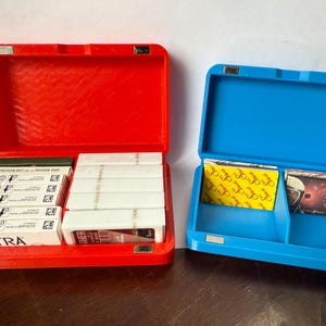 May include: Two open plastic cases, one red and one blue, each with a magnetic closure. The red case contains several packs of white and green boxes. The blue case holds yellow and brown boxes. The cases are on a brown surface.