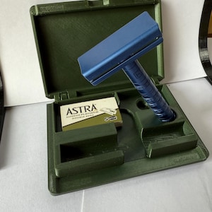 Puede incluir: Una maquinilla de afeitar de seguridad azul con un mango texturizado descansa en un estuche verde oscuro abierto. El estuche contiene un compartimento para la maquinilla y una ranura para un paquete de cuchillas de afeitar Astra Superior Platinum Double Edge.