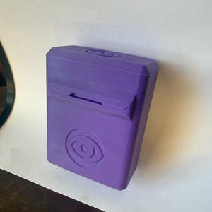 May include: A purple rectangular box with a hinged lid and a recessed opening. The box features a circular eye logo on the front. The box is made of a solid material and has a matte finish.