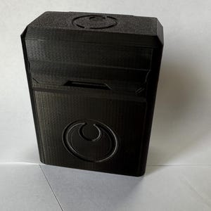 May include: A black, rectangular box with a textured surface. The box features a circular emblem on the front and a recessed design on the top. The box appears to be 3D-printed.