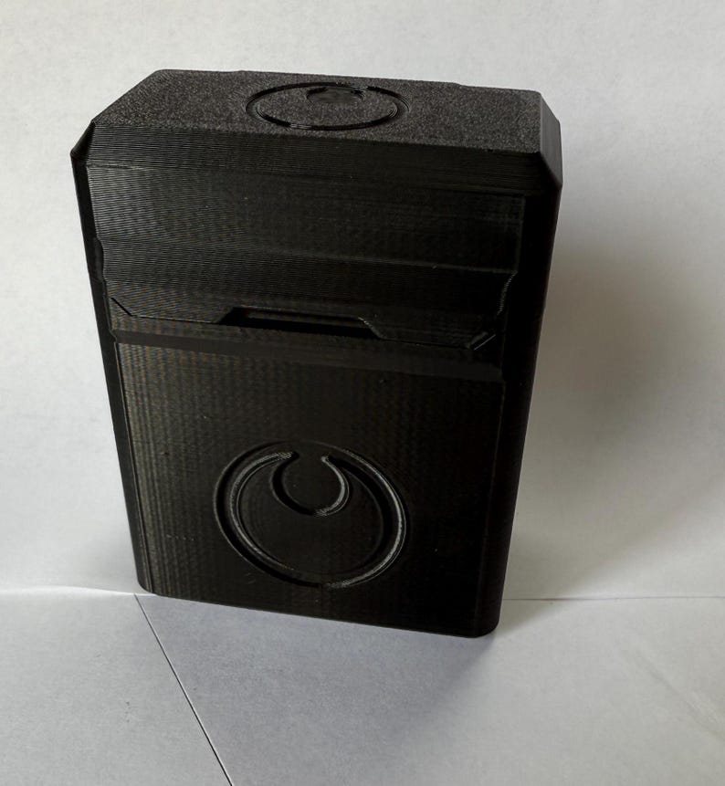 May include: A black, rectangular box with a textured surface. The box features a circular emblem on the front and a recessed design on the top. The box appears to be 3D-printed.