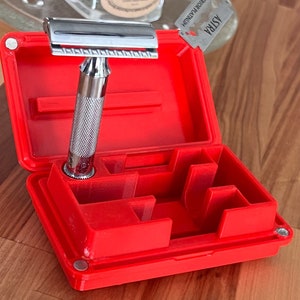 May include: A chrome safety razor with a textured handle sits in an open, bright red travel case. The case has compartments to hold the razor. The case has a magnetic closure. The razor is designed for shaving.