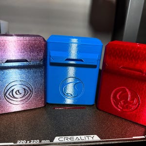 May include: Three 3D-printed rectangular cases in a row. The case on the left is a gradient of purple and blue with an eye symbol. The middle case is blue with a wave symbol. The case on the right is red with a symbol.