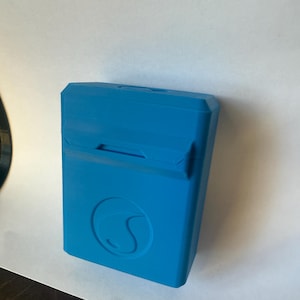 May include: A bright blue rectangular case with a curved design on the front. The case has a hinged lid with a small opening. The case is made of a solid material and appears to be 3D printed.