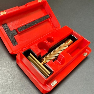 Puede incluir: Estuche rojo abierto con una maquinilla de afeitar dorada en su interior. La maquinilla tiene un mango texturizado y se guarda en un compartimento a medida. El estuche tiene un inserto de espuma negra y cierres magnéticos. Accesorio de afeitado clásico.