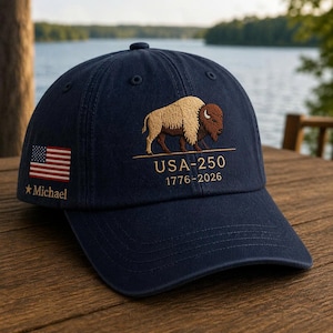 May include: Navy blue baseball cap with an embroidered bison and the text "USA-250 1776-2026." Features an American flag and the name "Michael" embroidered on the side. A classic, patriotic accessory.