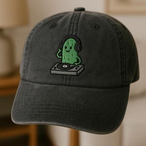 May include: A dark gray baseball cap featuring an embroidered green cactus wearing headphones and operating a turntable. The cap has a curved brim and a button on top, offering a casual and stylish accessory.
