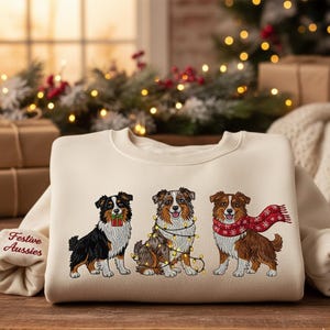 May include: Cream-colored sweatshirt featuring three embroidered Australian Shepherds. One dog holds a gift, another is adorned with Christmas lights, and the third wears a red scarf. The sleeve reads "Festive Aussies".
