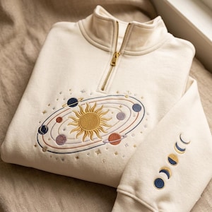 May include: Cream-colored sweatshirt with a gold zipper and embroidered celestial design. The design features a sun, planets, and stars on the front, with moon phases on the sleeve. The sweatshirt has a high collar.