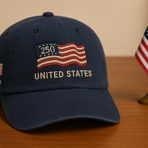 Op de afbeelding: Marineblauwe baseballcap met een geborduurd Amerikaans vlagpatch met de tekst "250 UNITED STATES". Aan de zijkant zit een kleine Amerikaanse vlag, met de naam "John" eronder geborduurd. Er is ook een miniatuur Amerikaanse vlag aanwezig.