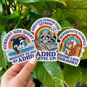 May include: Three embroidered patches with rainbow designs and text. One features a whale, another a raccoon, and the third a squirrel. The patches have text that reads "Current Side Quest", "Executive Dysfunction Extreme", and "Hyperfocus Olympics".