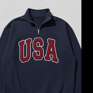 May include: Navy blue sweatshirt with a quarter-zip collar. The word "USA" is embroidered in white with a red outline across the chest. The sweatshirt has a casual, sporty style.