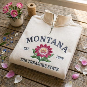 May include: A cream-coloured sweatshirt with a quarter-zip collar. The front displays the word "MONTANA" in navy blue embroidery above a pink and green floral design. Below the flower, the text reads "THE TREASURE STATE".