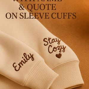May include: Cream-colored sweatshirt sleeves with embroidered text. One cuff reads "Emily" and the other reads "Stay Cozy" with a heart. The image also includes the text "Personalize with Name & Quote on Sleeve Cuffs".