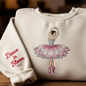 May include: Cream-colored sweatshirt featuring an embroidered ballerina with a pink and silver tutu. The sleeve displays the text "Dance in Bloom" in red script. A ballet-themed apparel item.