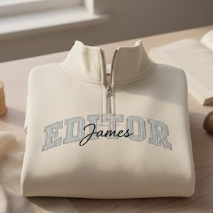 May include: A cream-colored sweatshirt with a quarter-zip collar. The word "EDITOR" is embroidered in a light blue and purple collegiate font, with the name "James" embroidered below in a cursive font. The sweatshirt is folded on a wooden surface.