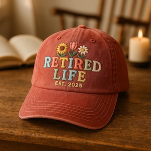 May include: A red baseball cap with the embroidered text "RETIRED LIFE EST. 2025" in colorful letters. The cap features embroidered flowers above the text, including a sunflower, tulip, and daisy. The hat is made of a soft, worn material.