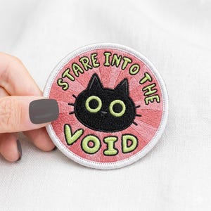 May include: Embroidered circular patch featuring a black cat face with green eyes and the text "STARE INTO THE VOID" in green and pink. The patch is set against a white backdrop.