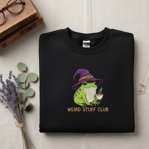 May include: Black sweatshirt featuring an embroidered green frog wearing a purple witch hat and holding a coffee cup. The text "WEIRD STUFF CLUB" is embroidered in orange below the frog. The sweatshirt is on a neutral surface.