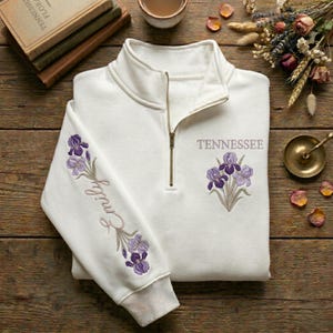 May include: A white quarter-zip sweatshirt with embroidered purple iris flowers and the word "Tennessee." The left sleeve features the name "Emily" in script. The sweatshirt has a ribbed collar and cuffs, and a gold zipper.
