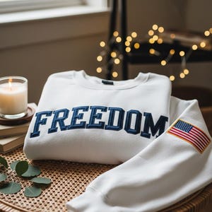 May include: A white sweatshirt with the word "FREEDOM" embroidered in navy blue across the chest. The sleeve features a patch of the American flag. The sweatshirt is folded on a woven table, with a lit candle and books in the background.