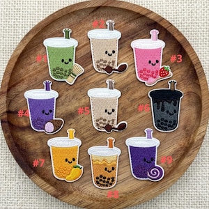 May include: A collection of nine embroidered patches featuring bubble tea designs. Each patch showcases a different color and flavor, including green, brown, pink, purple, yellow, and black. Some patches include additional elements like a strawberry or a mango.