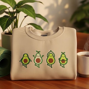 May include: Beige sweatshirt featuring embroidered avocados in yoga poses. The avocados are green with brown centers and smiling faces. A rolled-up green yoga mat and a cup of coffee are in the background.