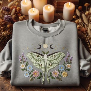May include: A light gray sweatshirt with a crew neck featuring an embroidered design of a moth, crescent moons, and flowers. The moth is light green with detailed wing patterns. Floral accents include blue, pink, and yellow flowers.