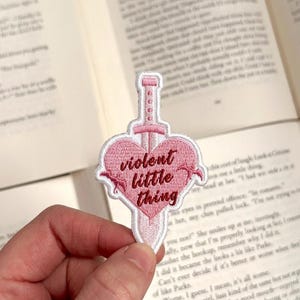 May include: Embroidered patch featuring a pink heart pierced by a sword. The patch reads "violent little thing" in a cursive font. The patch is held in front of an open book.