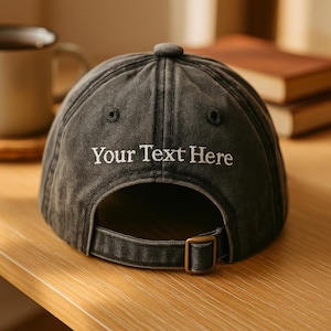 Op de afbeelding: Een antracietgrijze baseballpet met de tekst "Your Text Here" in het wit geborduurd. De pet heeft een gebogen klep, een metalen gespsluiting en een kleine knoop aan de bovenkant. De hoed is gemaakt van zacht, versleten materiaal.