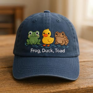 May include: Navy blue baseball cap with embroidered cartoon frog, duck, and toad. The text "Frog, Duck, Toad" is embroidered below the characters. The cap is made of a soft material and has a curved brim.