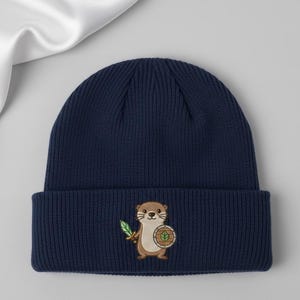May include: Navy blue knit beanie featuring an embroidered cartoon otter holding a sword and shield. The otter is brown and tan with a white face and whiskers. The shield is green and brown.